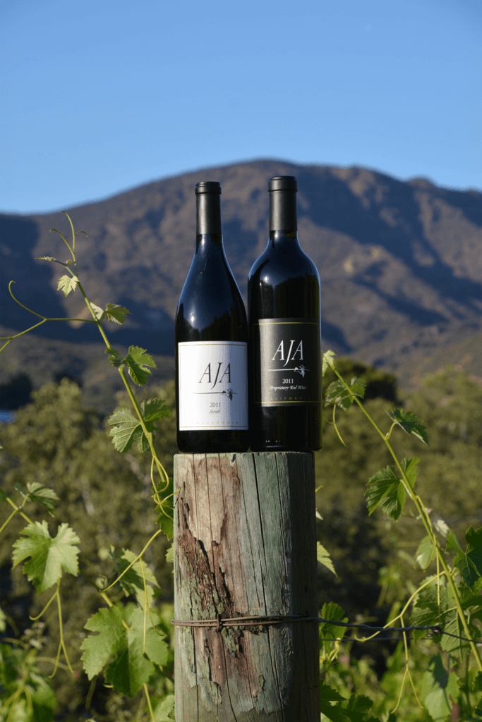 Two bottles of AJA wine standing on a vineyard post with the Santa Monica Mountains behind